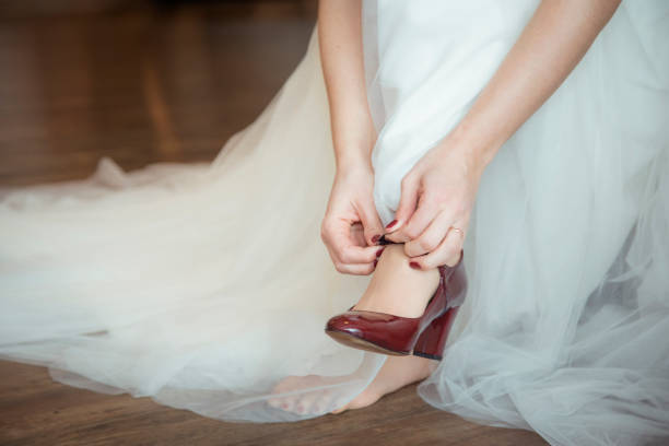 A bride is getting ready for her wedding, carefully fastening her red high-heeled shoes. Her elegant white gown flows around her as she prepares to walk down the aisle.