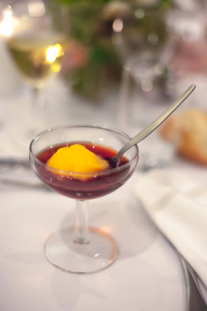 A delicate dessert features rich red sauce topped with a scoop of peach sorbet in a stylish glass. This delightful treat is served on a beautifully arranged dining table.