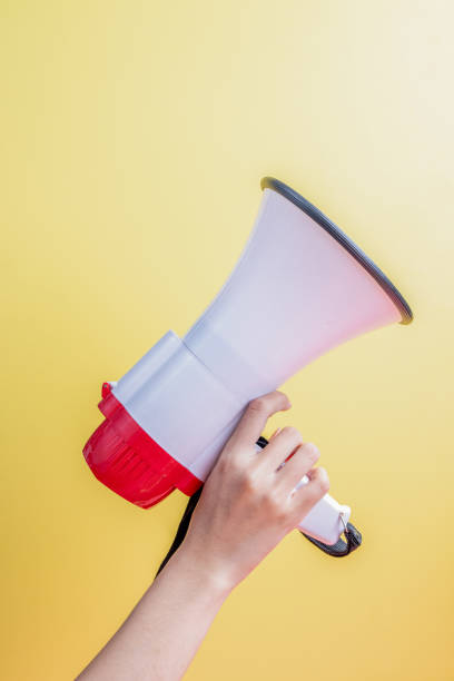 A hand holding a white and red megaphone against a bright yellow background, symbolizing communication, announcement, or activism. Ideal for themes related to marketing, public speaking