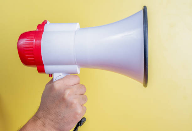 A hand holding a white and red megaphone against a bright yellow background, symbolizing communication, announcement, or activism. Ideal for themes related to marketing, public speaking