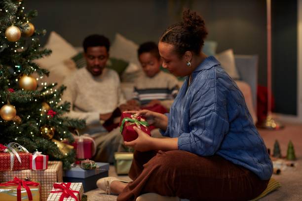 istock Smiling Black Woman Looking at Gifts Under Christmas Tree 2183012759