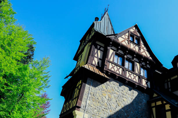 Brasov, Romania - May 23, 2023: Pelisor Castle showcases its captivating design with a blend of stone and wood nestled among lush green trees under a brilliant blue sky evoking a sense of history and natural beauty.