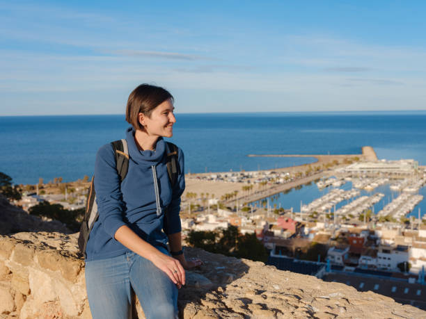 Young woman exploring historic castle in Denia, Alicante province, Spain, scenic views of town below, set against cooler season backdrop. blend of history and tranquility in timeless setting.