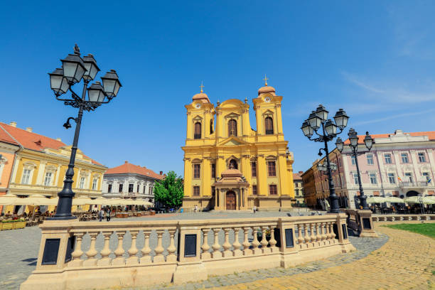 The bustling Union Square in Timisoara features a bright yellow church against a clear blue sky. People enjoy the lively atmosphere with outdoor dining and historical architecture.