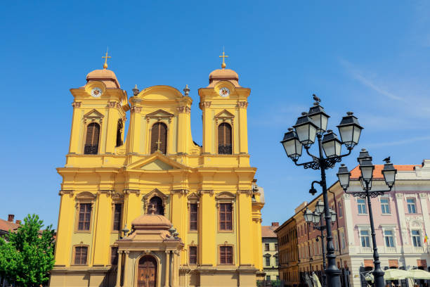 The vibrant yellow cathedral stands majestically under a clear blue sky in Timisoara Romania. Historic buildings and ornate street lamps frame this stunning architectural landmark.
