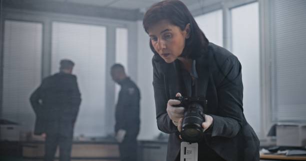 Office robbery or murder case: Female forensic scientist takes photos of evidence with camera. African American policeman and detective in the background. Investigation team working at crime scene.