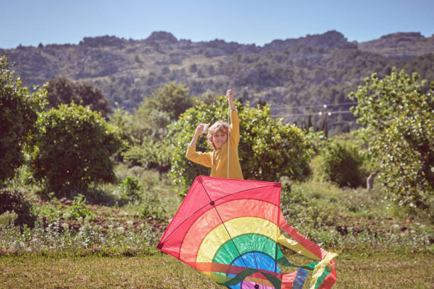 Cheerful boy pulling fallen on grass kite in summer day on green light lawn