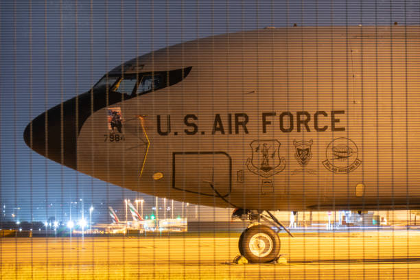The forward fuselage of a United States Air Force Boeing KC-135R Stratotanker plane, registration 63-7984, parked at Sydney Kingsford Smith Airport.  In the foreground is the airport's steel security fence. In the far background are the vertical stabilisers of two Emirates A380-861 and A380-842 planes, registrations A6-EEF and A6-EVL. This image was achieved with a 13 second exposure and taken from Ross Smith Avenue, Mascot, on a cloudy evening after sunset on 1 November 2024.