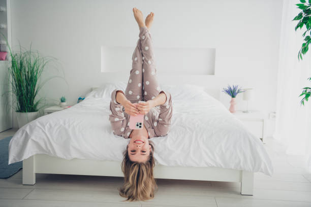 istock Young woman in polka dot pajamas lying upside down on cozy bed while using smartphone in sunlit bedroom 2183050989