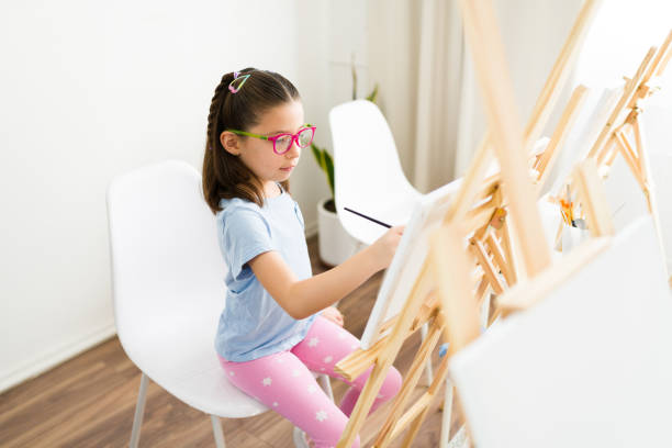 Cute girl with glasses creating a painting on a small canvas in an art class, nurturing her creativity and honing her artistic abilities at school