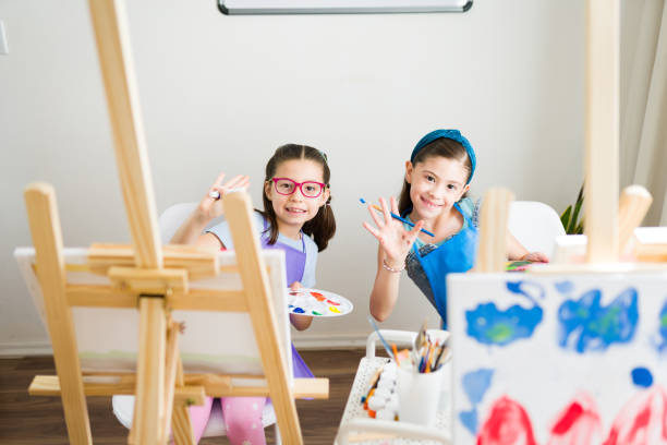 istock School painting class: two young students having fun and waving and saying hello while holding their paintbrushes 2183053299