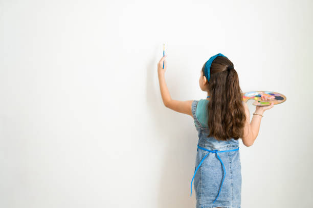Young elementary school student expressing creativity by painting on a blank wall in art class, holding a palette and brush