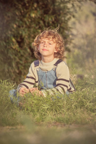 Adorable boy in casual clothes sitting on grassy shore and looking at camera