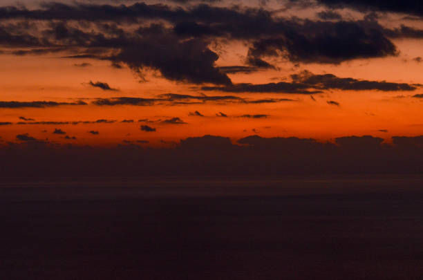 Experience the breathtaking view of a dramatic sunset over the sea, as dark clouds silhouette against an intense orange horizon.