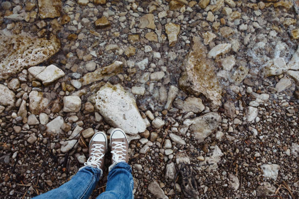 A high angle shot of a woman's shoes as she stands at the water's edge on a rocky beach.