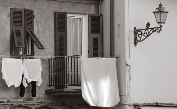 A charming Ligurian balcony scene with open shutters, a small wrought-iron railing, and laundry hanging out to dry. The black-and-white tones give the image a timeless feel, highlighting the textures of the wall, shutters, and hanging clothes. A vintage-style street lamp with a perched bird adds a nostalgic touch, evoking the quiet simplicity of Italian village life.