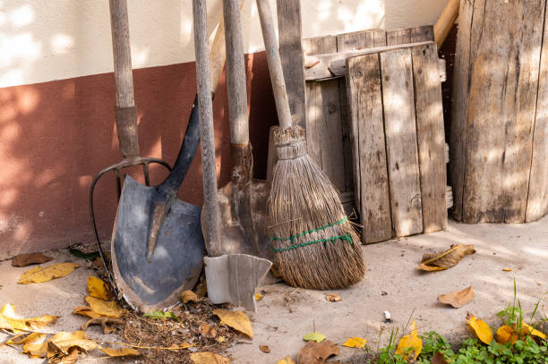 A selection of rustic gardening tools, including shovels, a broom, and a pitchfork, leaning against an outdoor wall with a wooden pallet nearby. Fallen autumn leaves add a seasonal touch to the scene, emphasizing a rural, traditional setting.