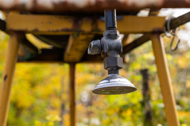 A close-up of a rustic garden shower faucet attached to a metal structure supporting a barrel, designed as an outdoor shower setup. The weathered metal and blurred natural background with autumn hues evoke a rural, handmade aesthetic.