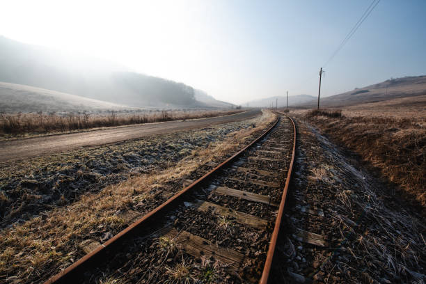 A misty winter scene featuring a desolate road running parallel to rusty railway tracks through a frosty, barren landscape. The poles and distant hills add to the quiet, solitary atmosphere of the countryside in winter.