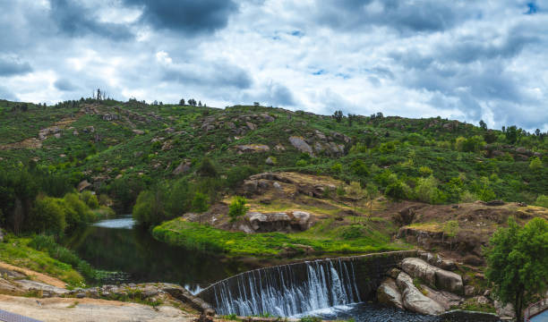 A serene landscape featuring a gentle waterfall flowing over a stone dam, surrounded by lush green hills under a moody, cloudy sky