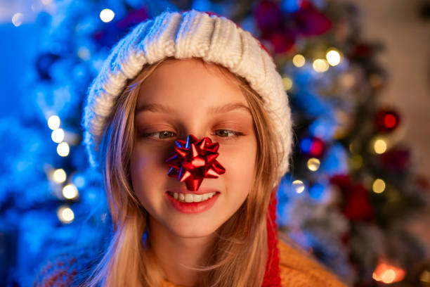 Young girl playfully wearing a red Christmas bow on her nose, smiling in front of a decorated Christmas tree during the holidays.