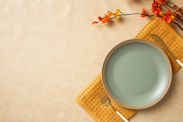 Minimalist autumn table setting with green plate, gold fork and knife, branch with orange leaves on beige background. Ideal for fall themes, seasonal decor, and cozy dining