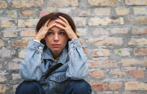 istock Woman with a tired, worried expression sits outdoors next to a brick wall, hands on her forehead. 2183128717
