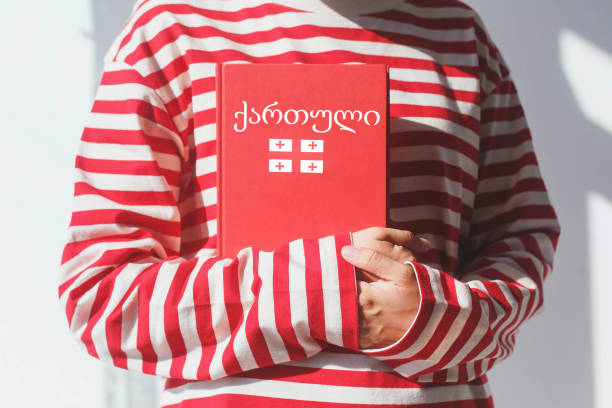 istock One girl, whose face is not visible in a striped red long sleeve, holds a red Georgian language textbook with Georgian flag and Georgian inscription in Georgian, foreign language learning concept 2183137826