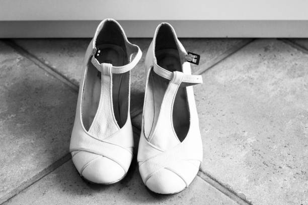 A pair of stylish white T-strap heels rests on a textured tiled floor. The soft natural light highlights the shoes' elegant design in a contemporary indoor space.