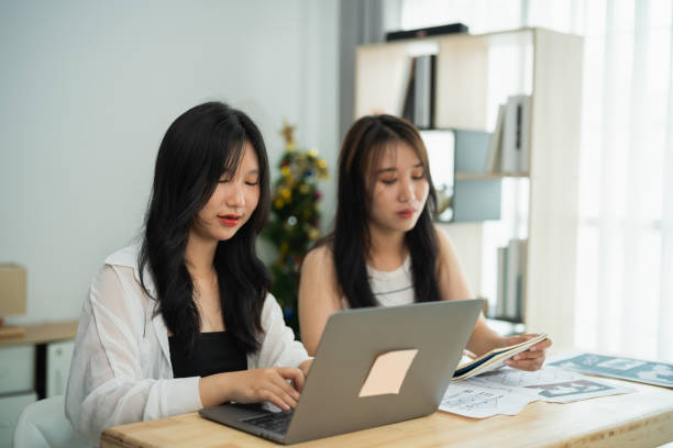 istock Two Asian women are sitting at a table with a laptop and a stack of papers. They are working together on a project 2183182821