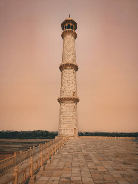 This striking image captures a tall, cylindrical minaret made of white marble, standing on a large, open, tiled platform. The minaret has multiple balconies and a domed top. The platform is bordered by a low railing, and there is a door at the base of the minaret. The background features a hazy sky and a distant tree line. This image is interesting because it captures the architectural beauty and intricate design of the minaret, which is a significant element in many historical and cultural sites.
