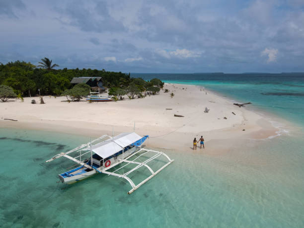 A couple strolls along the white sandy shores of a tropical island, surrounded by turquoise waters and lush greenery. A boat rests nearby, enhancing the peaceful island ambiance.
