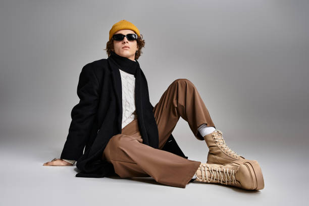 A stylish young man poses in a black coat and yellow beanie against a backdrop