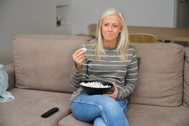 istock Senior woman enjoying popcorn while watching tv on sofa at home 2183226890