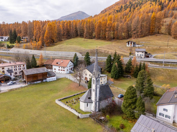 A picturesque Alpine village with charming houses and a tall church steeple, surrounded by vibrant autumn trees and rolling hills under a bright sky. Tranquil and timeless beauty.