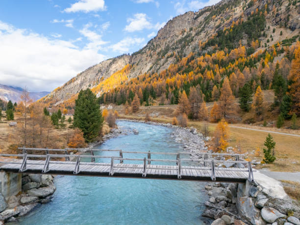 A wooden bridge spans a crystal-clear river in a picturesque autumn valley, framed by vibrant fall foliage and rugged mountain slopes under a partly cloudy sky.