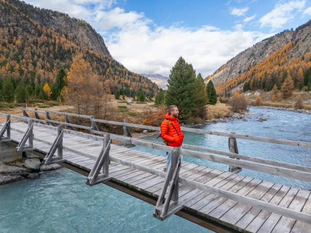 A person wearing a red jacket stands on a wooden bridge over a vibrant blue river in an autumn landscape surrounded by mountains and colorful trees.