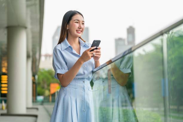 istock Young Woman Holding Phone Outdoors with Cityscape and Greenery Background, Looking Away Contentedly 2183244621