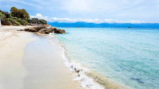 A quiet, sandy beach on Corsica with a stretch of clear, blue sea bordered by rocky formations. The beach is mostly empty, creating a peaceful, secluded atmosphere. The gentle waves lap at the sand, blending the coastal elements of sand, rocks, and water under a bright sky.