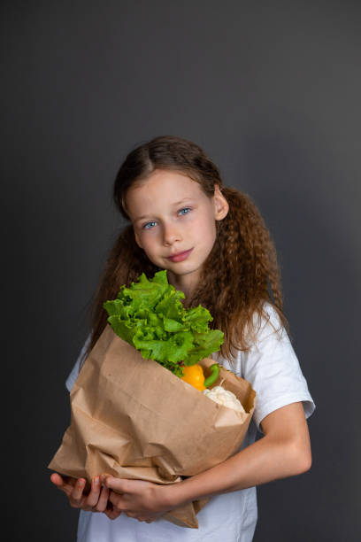 A girl stands in a white T-shirt holding a bag of vegetables and smiling on a gray background. Healthy eating children. Recipes and proper nutrition for a child. Proper nutrition for teenagers. Vitamins and nutrients in food for children. Childhood obesity. Nutritionology. Diet. Poor nutrition in teenagers una niña está de pie con una camiseta blanca sosteniendo una bolsa de verduras y sonriendo sobre un fondo gris. alimentación saludable para niños. vitaminas y nutrientes en los alimentos para niños. obesidad infantil. nutriología. dieta. mala nutrici