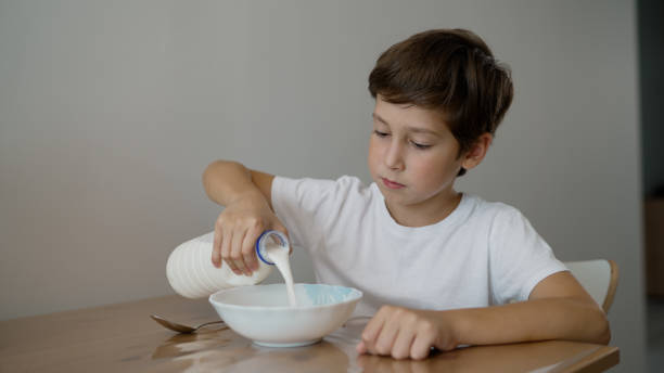 istock Healthy breakfast for a child. Proper nutrition, children's menu. The boy eats cereal for breakfast. A boy adds milk to porridge while sitting at the table. A brunette in a white T-shirt is eating at the table. 2183253225