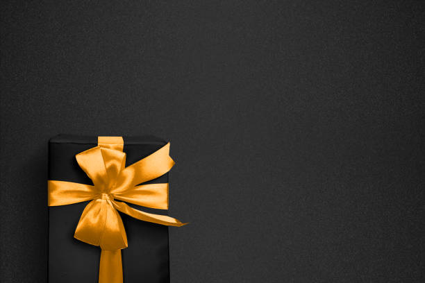 Black gift box decorated with a textured golden bow on black background. Copy space. Mockup or template. Birthday, anniversary presents, gift post cards. Flat lay