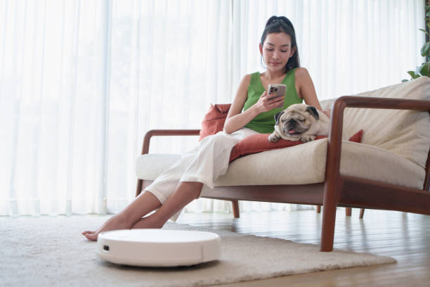 istock Young Beautiful Asian woman relaxing on sofa with her pug dog while using mobile phone or smartphone in living room while robotic vacuum working on floor. 2183270874
