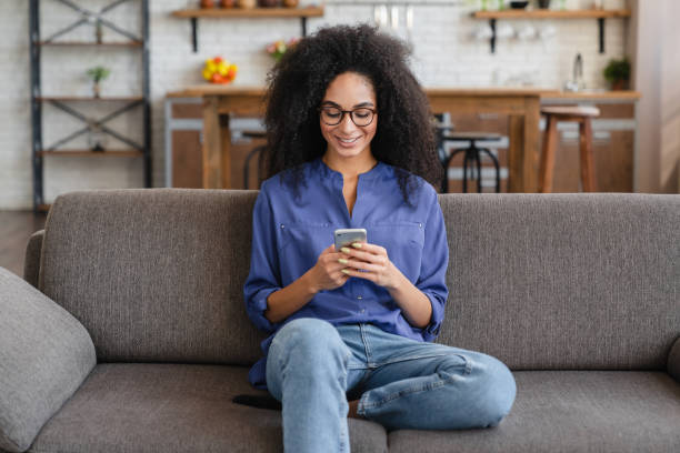 istock Smiling young African woman using cell phone relaxing on couch at home. 2183306990