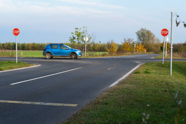 A blue car drives through a rural intersection with multiple stop signs on a clear autumn day. The scene captures the open countryside with green fields and seasonal trees in the background.