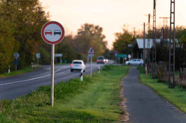 A no overtaking sign stands beside a rural road as vehicles drive into the distance at sunset. The soft evening light and surrounding greenery create a calm and scenic countryside atmosphere.