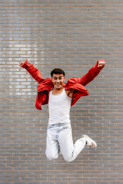 Latin LGBT young man with red overshirt jumping with joy in front of a brick wall