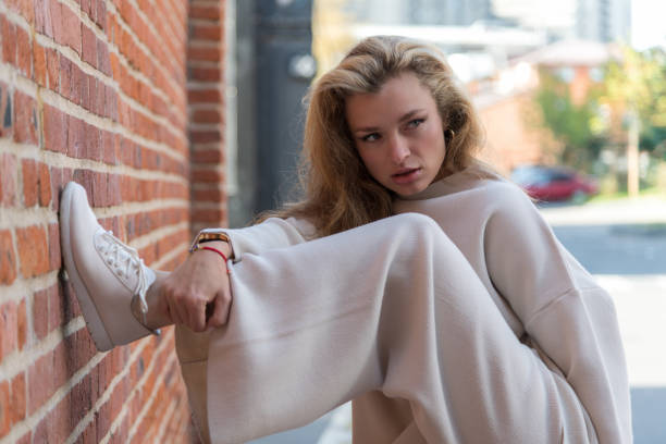 Close-up side view portrait of beautiful long haired serene caucasian blond woman in white sweater standing on one leg by red brick wall on city street in autumn day. Soft focus. Fashion theme.