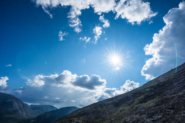 A stunning view of a bright sunlit sky shining over a rugged mountain landscape, accented by fluffy clouds. The scene evokes feelings of tranquility and awe-inspiring nature.