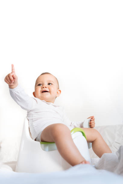 A delightful image of a baby sitting on a potty and pointing upward with a curious and joyful expression. Dressed in white, the baby radiates innocence and wonder, with soft natural light illuminating the scene. This photo is perfect for themes related to early learning, child development, and baby milestones.